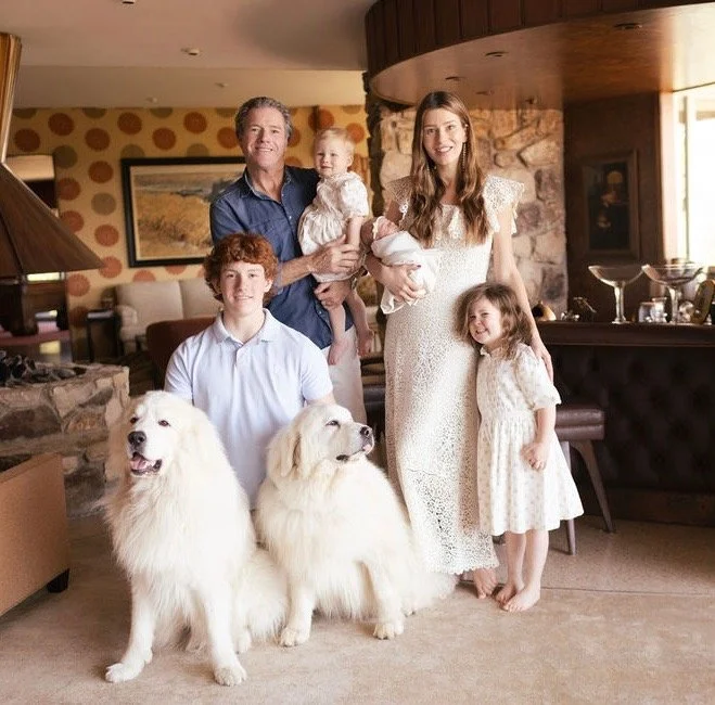 Family of five with two dogs in a cozy living room, including a man, a woman, three children, and two large white dogs.
