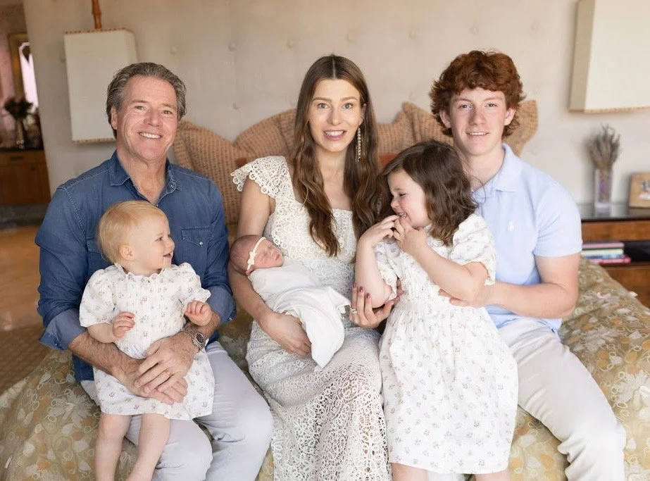 Family sitting on a bed, with a newborn and young children, smiling at the camera in a cozy bedroom.