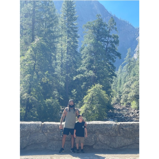 A man and a boy standing in front of a stone barrier with a forest and mountain background during daytime.