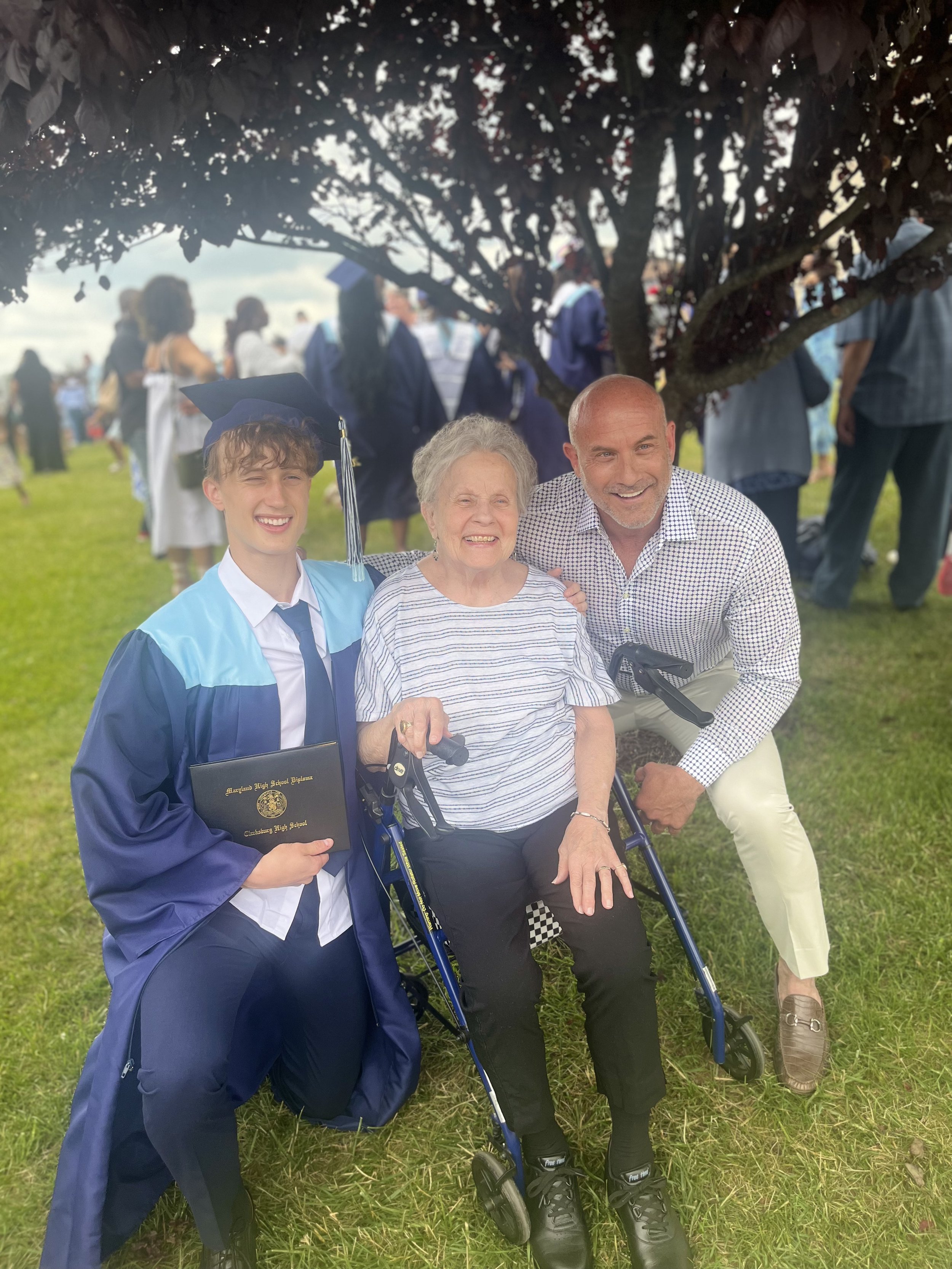 A young man in a graduation gown and cap with a diploma, an elderly woman in a wheelchair, and a middle-aged man, all smiling, at a graduation ceremony outdoors under a tree.