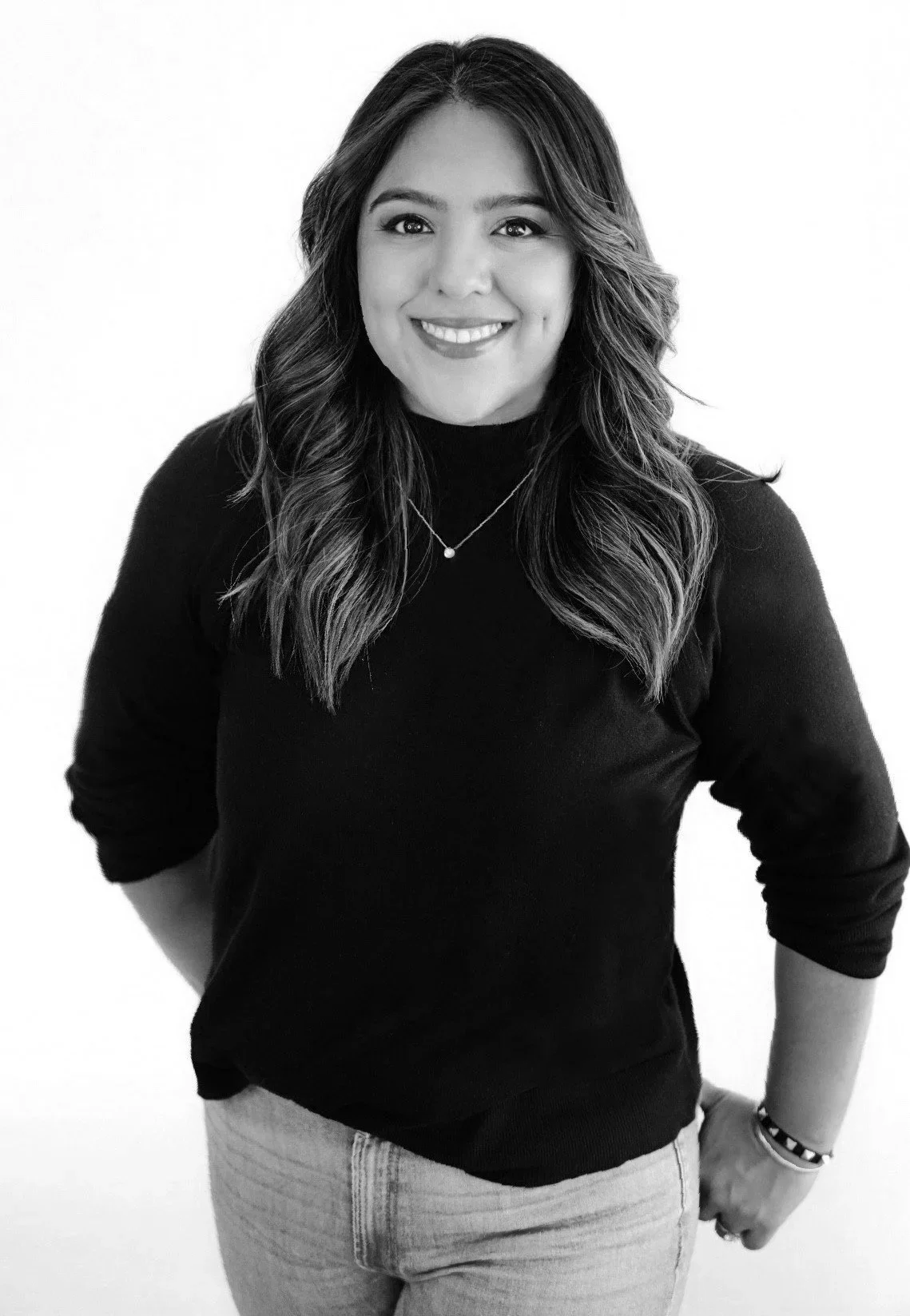 Black and white portrait of a young woman with wavy hair, wearing a black sweater, jeans, and a necklace, smiling at the camera.