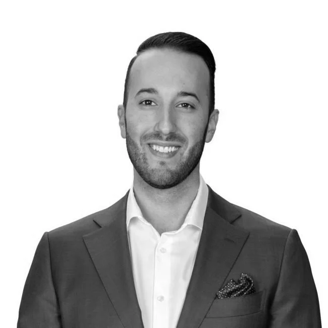 Black and white portrait of a smiling man with dark hair, wearing a suit jacket over a white dress shirt, with a pocket square.