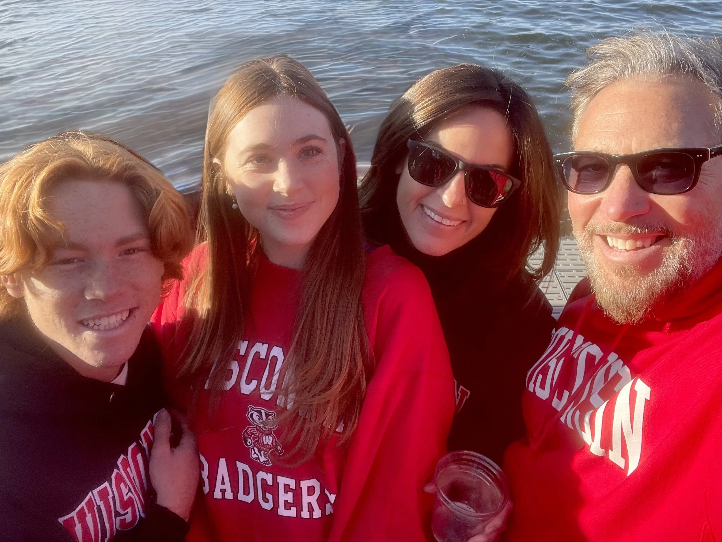 Four people smiling for a selfie near a body of water, wearing Wisconsin Badgers apparel.