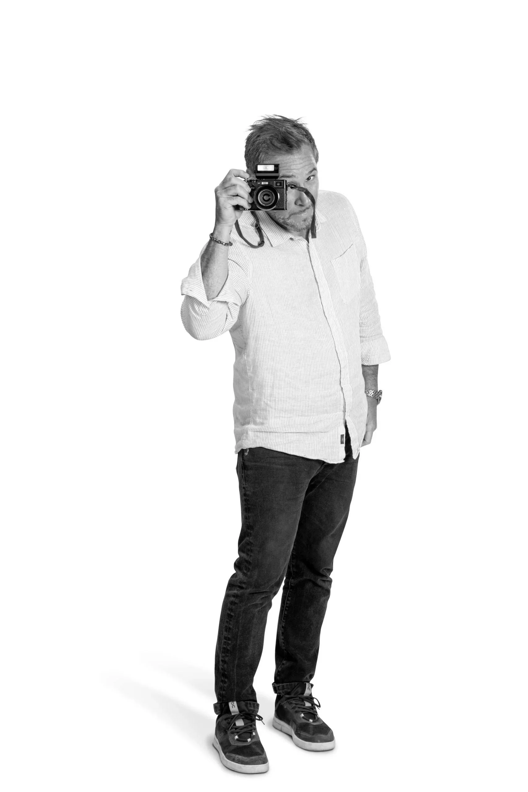 A man stands against a white background, holding a camera up to his eye and taking a photo.