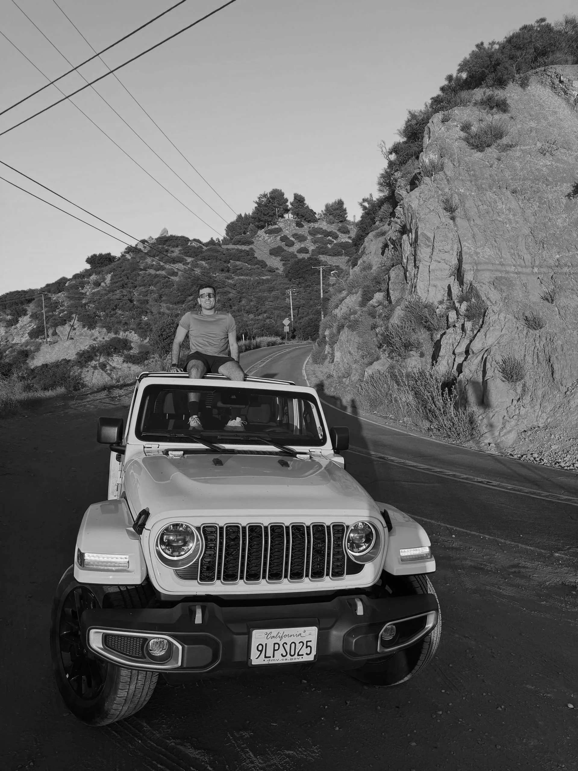 A person sitting on the roof of a Jeep Wrangler on a winding mountain road with hills and rocks in the background, black-and-white photo.