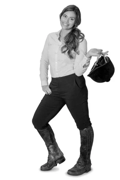 A woman standing and smiling, holding a black hat in her right hand, dressed in a white shirt, dark pants, and boots, in a black-and-white photo.