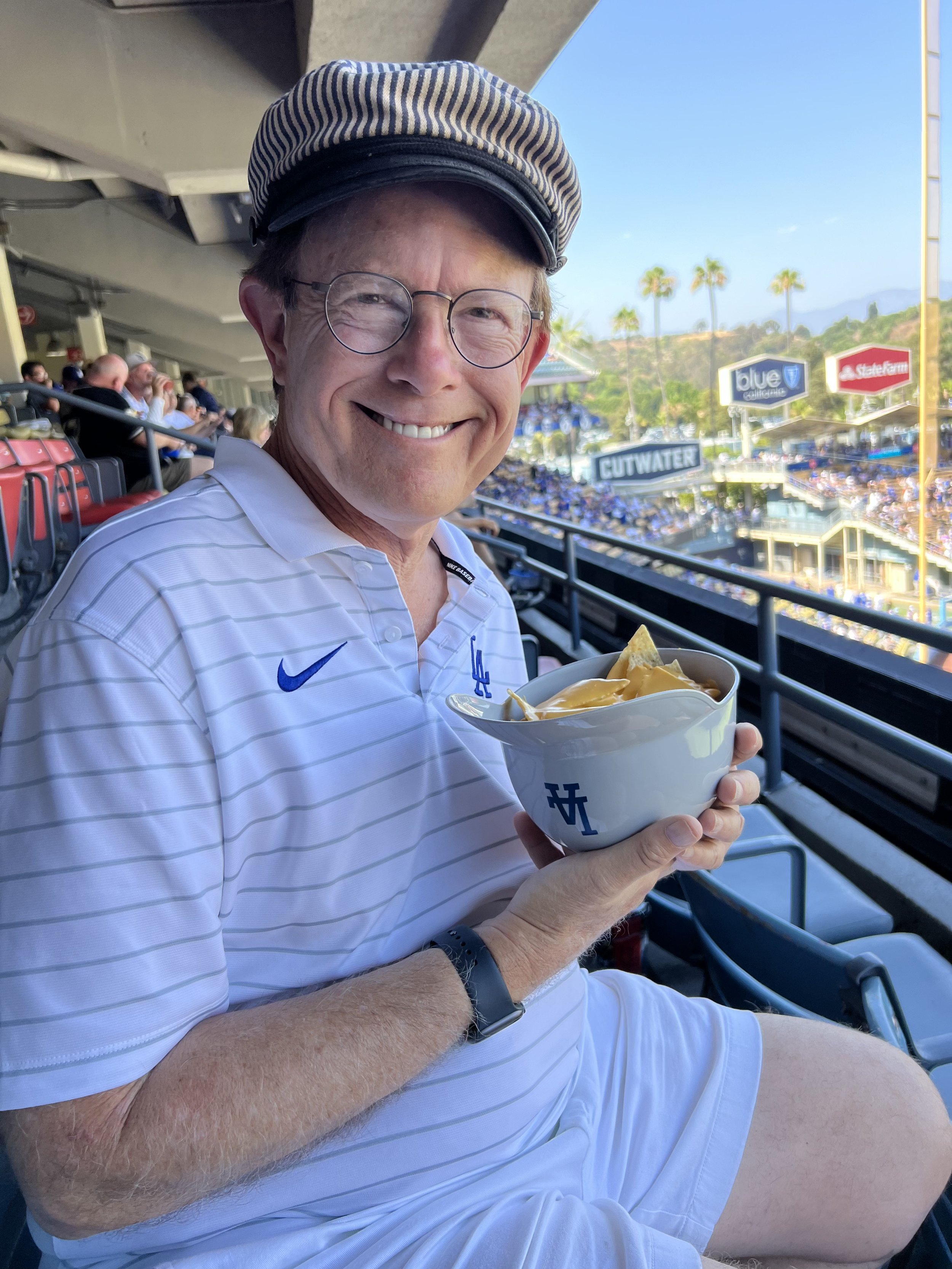 A man smiling at a baseball game, wearing a Los Angeles Dodgers shirt and hat, holding a bowl of nachos with cheese, with a crowded stadium and palm trees in the background.
