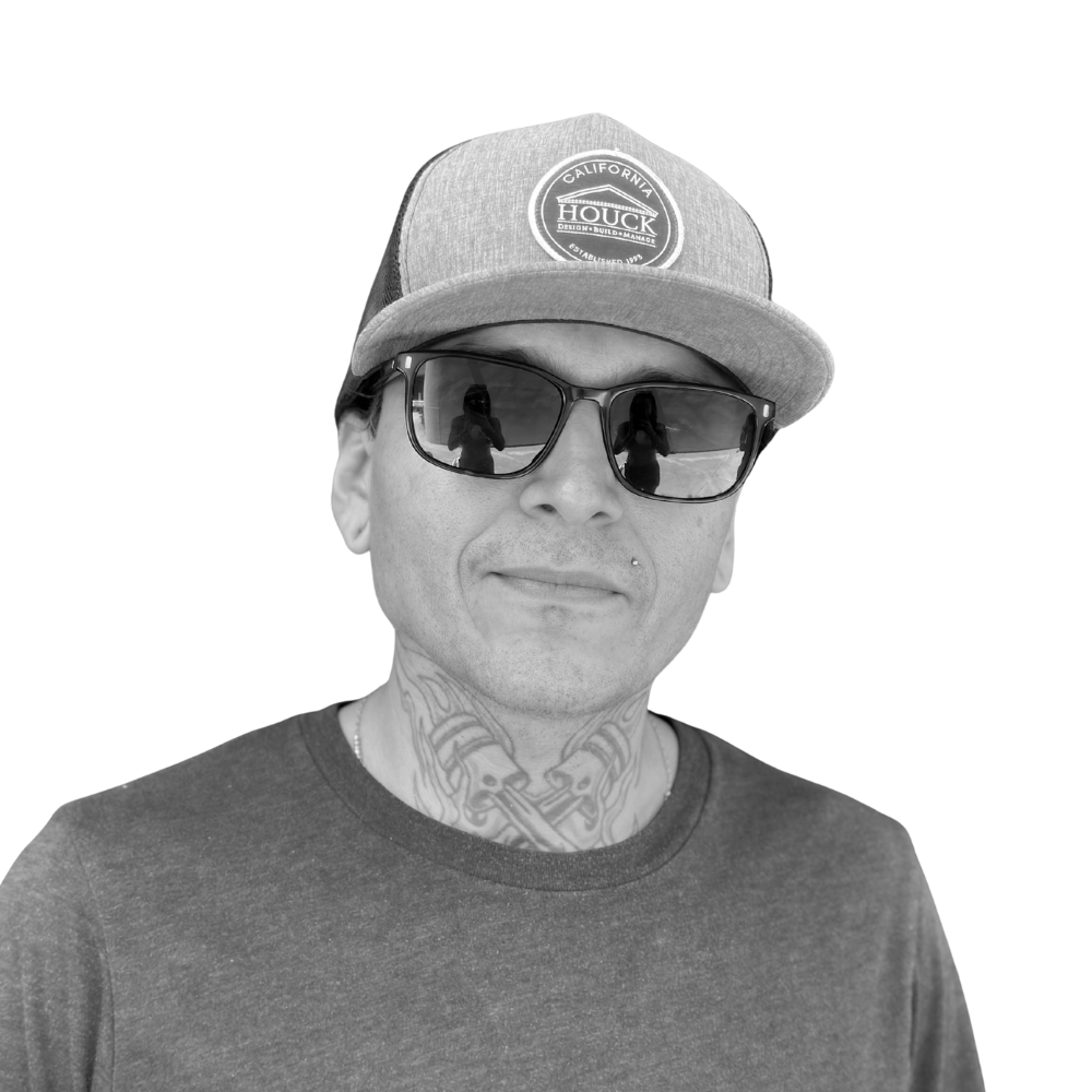 A man wearing a baseball cap, sunglasses, a t-shirt, and tattoos on his neck, smiling against a white background.