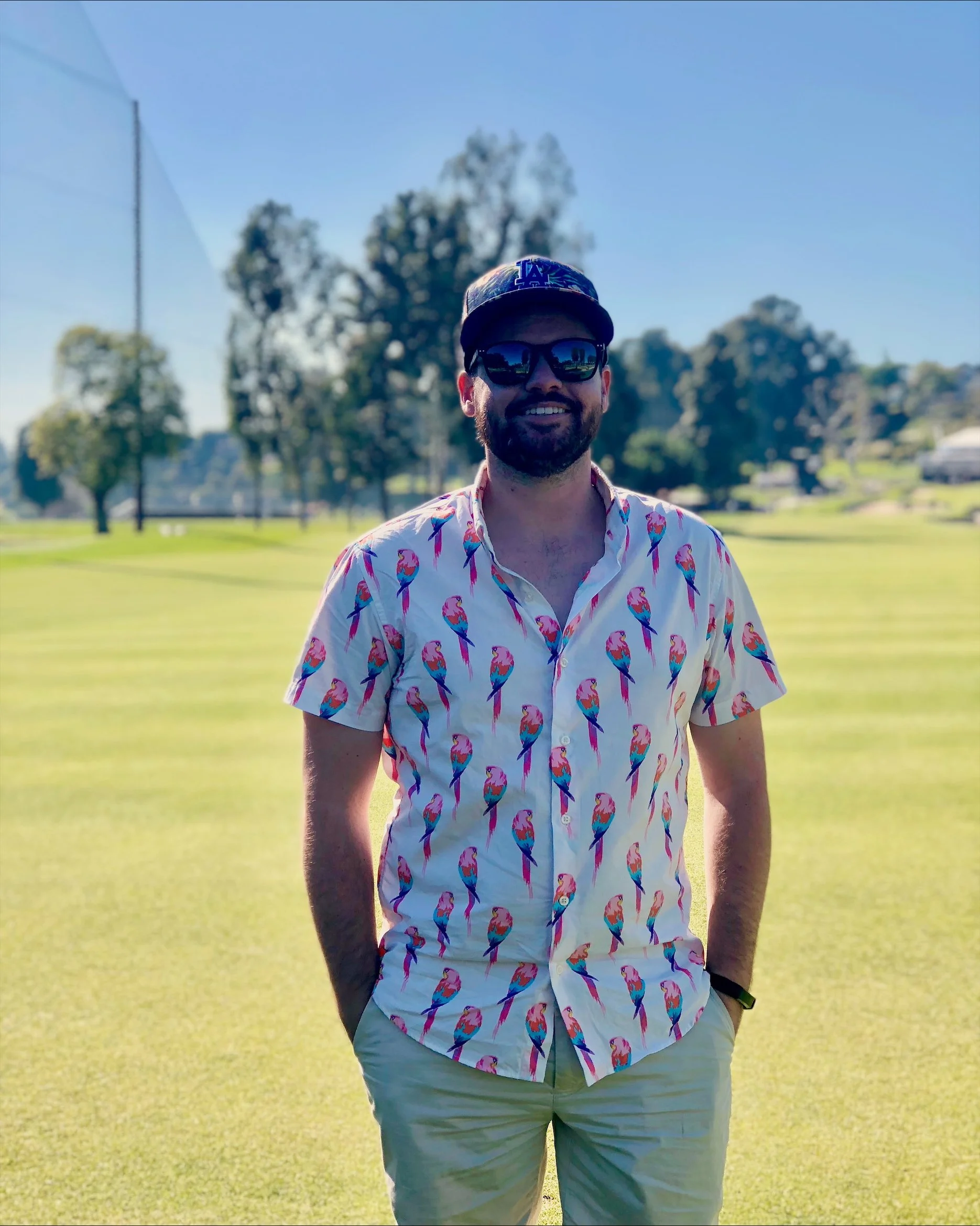 A man standing on a grassy field, wearing a colorful short-sleeved shirt with a flamingo pattern, beige pants, a dark cap, sunglasses, and a watch, smiling outside on a sunny day.