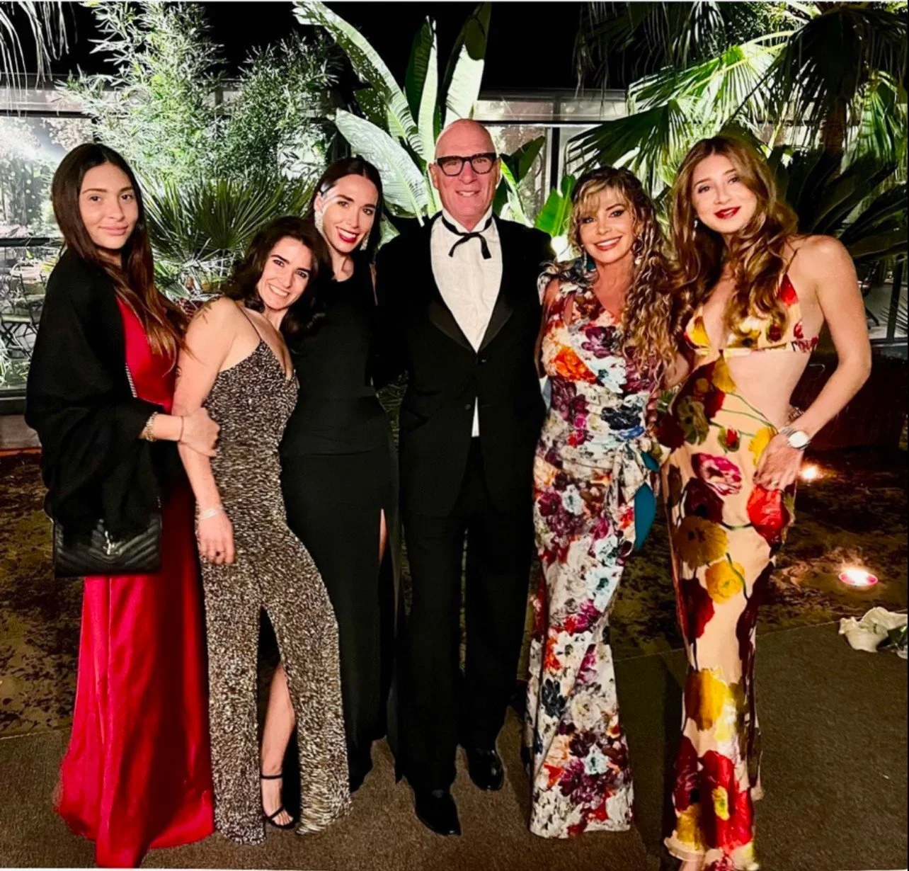 A group of six women and one man in formal attire posing together in a lush, indoor garden setting with greenery and decorative plants in the background.