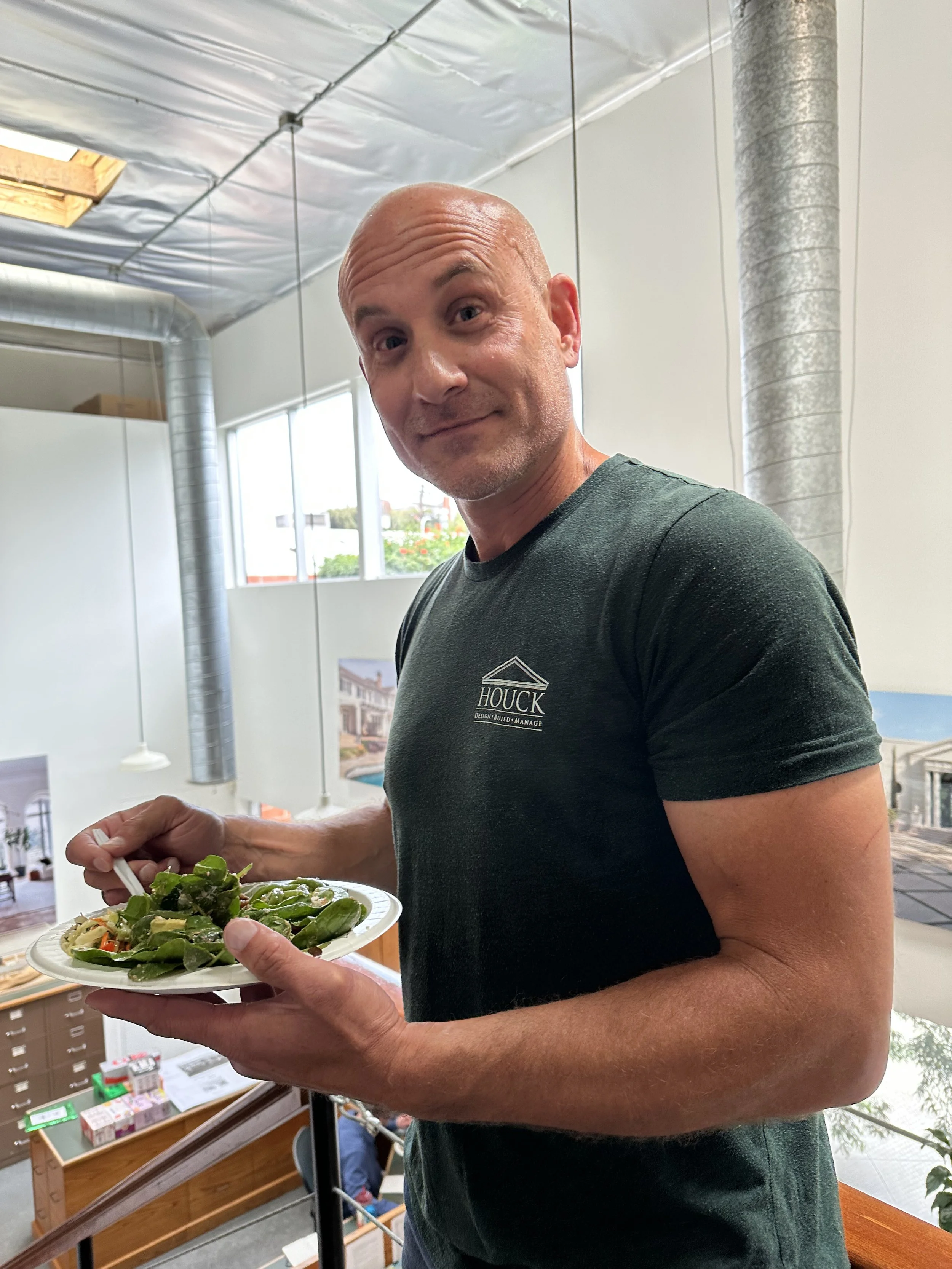 A man with a bald head and wearing a black T-shirt is holding a white plate of salad, standing inside a bright room with large windows and ductwork on the ceiling.