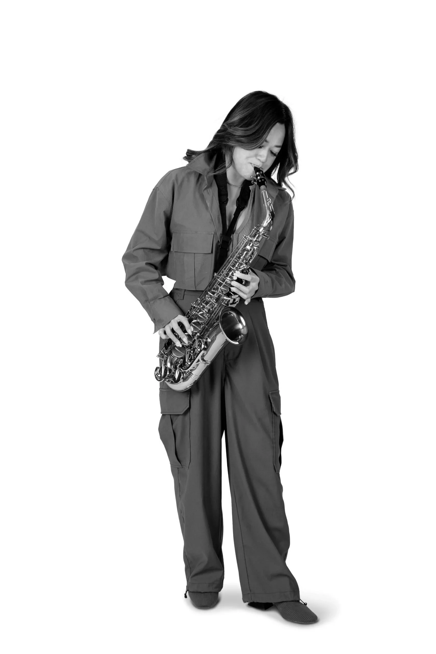 A woman playing a saxophone, dressed in a jacket and cargo pants, standing against a plain white background.