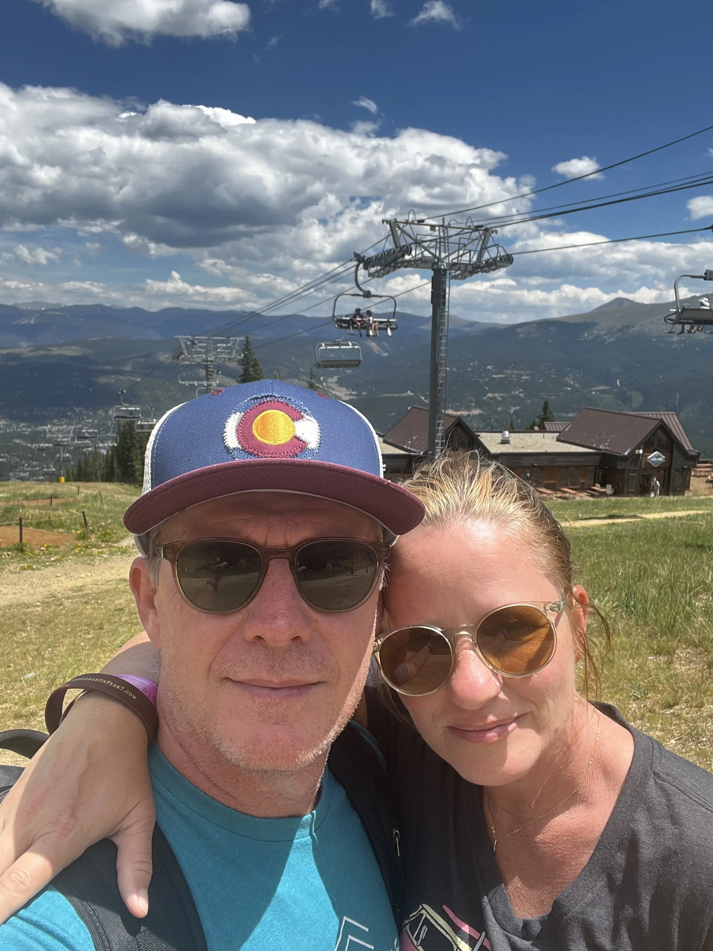 A middle-aged couple wearing sunglasses, smiling at the camera while taking a selfie outdoors. The man is wearing a Colorado cap and a blue shirt, and the woman has her arm around his shoulder. Behind them is a grassy area, ski lifts, and a building with mountains in the distance under a partly cloudy sky.
