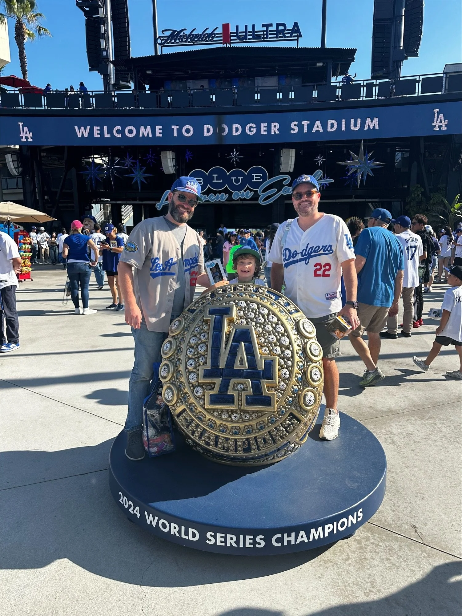 Three fans of the Los Angeles Dodgers pose with a decorative giant baseball with the Dodgers logo, celebrating their 2024 World Series championship outside Dodger Stadium with a crowd in the background.