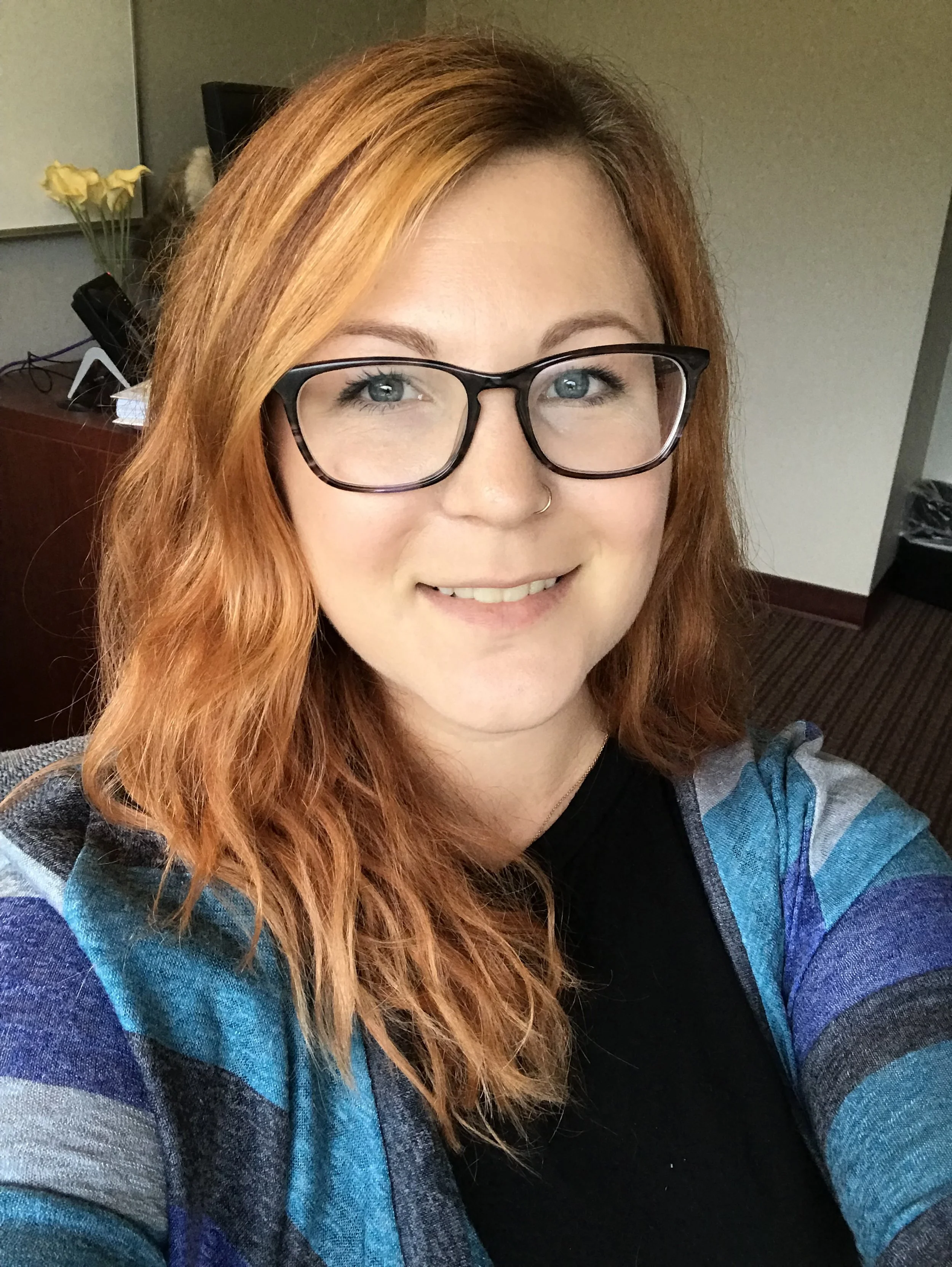 A woman with long, curly red hair, glasses, and a nose ring smiling in an indoor setting.