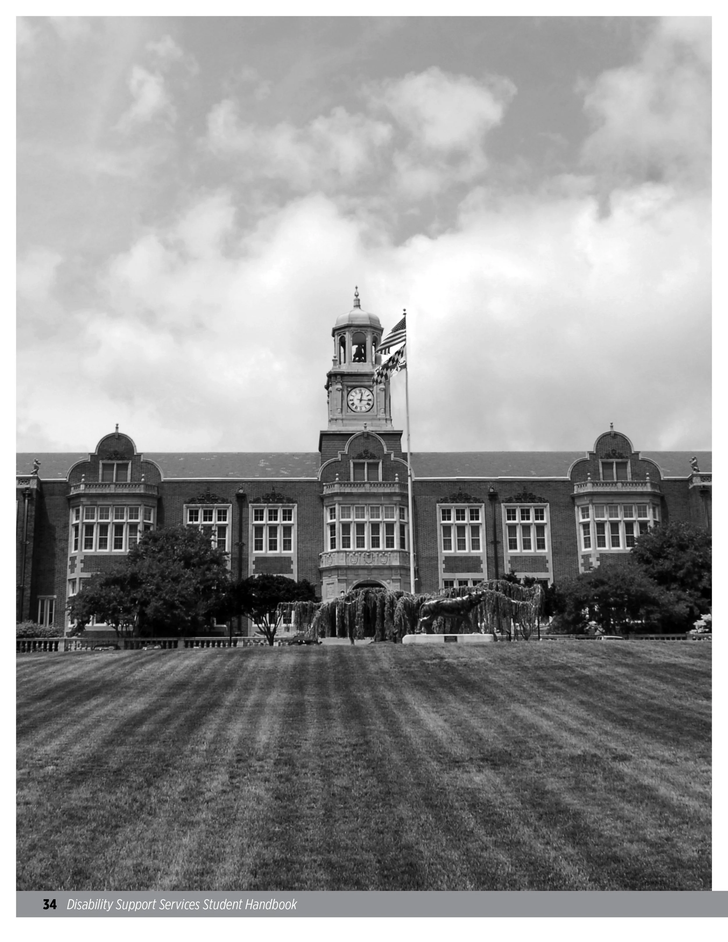 Black and white photo of a large historic building with a clock tower, American flags, and a manicured lawn with trees and a statue of a tiger in the foreground.