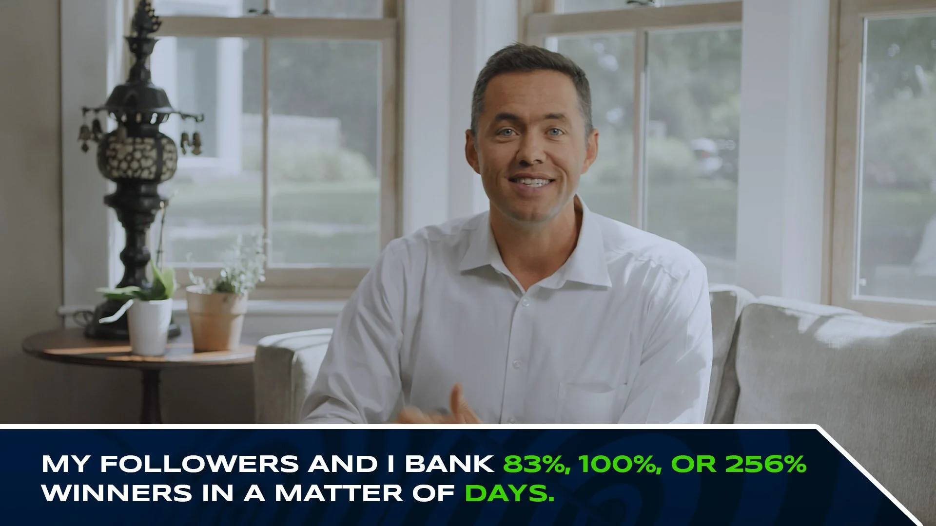 A man in a white shirt sitting on a sofa, smiling at the camera, with a window and potted plants behind him.