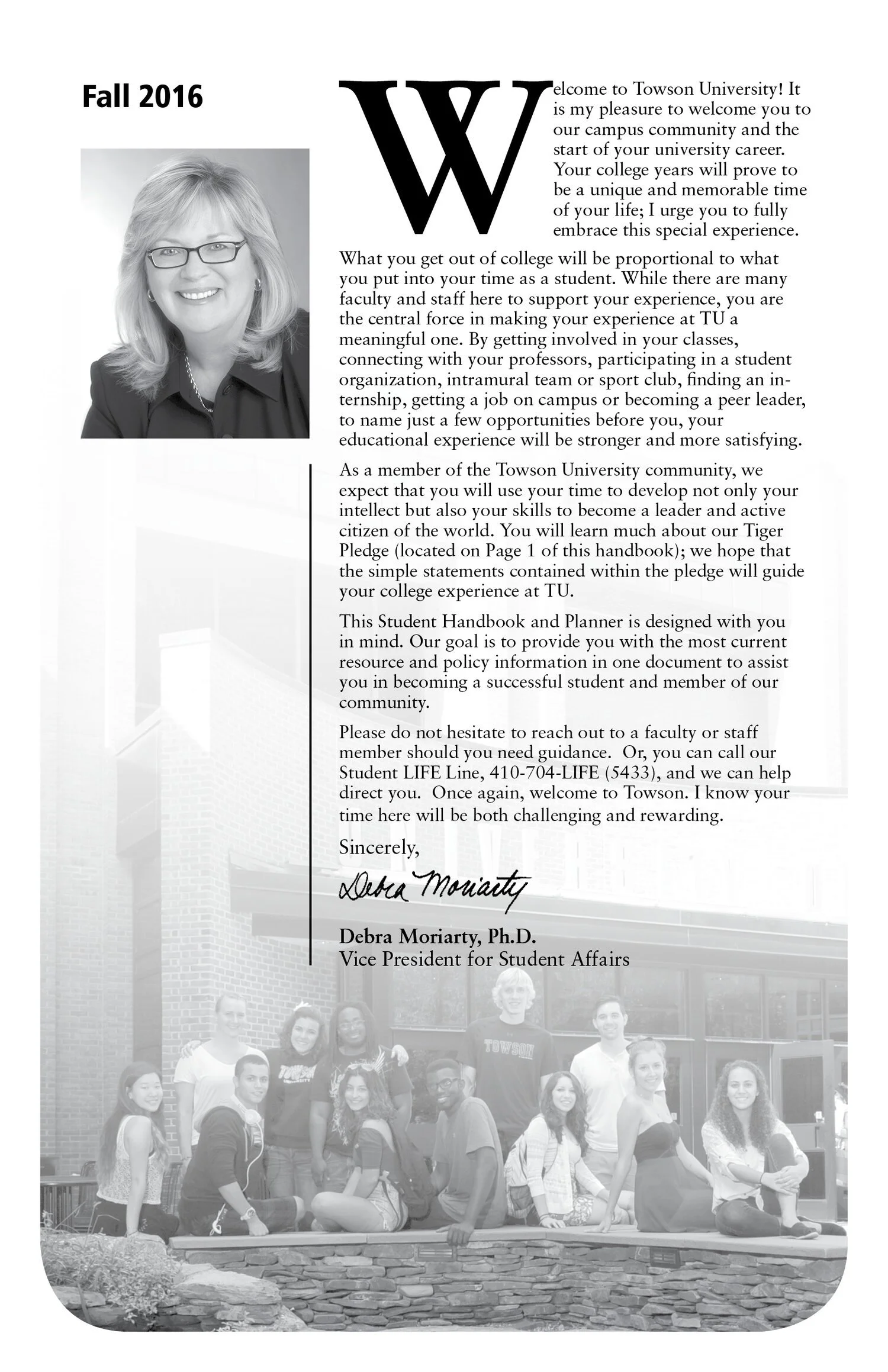 A black and white photograph showing a group of students sitting and standing outdoors in front of Towson University, with a brick building in the background, surrounding a lawn with plants.