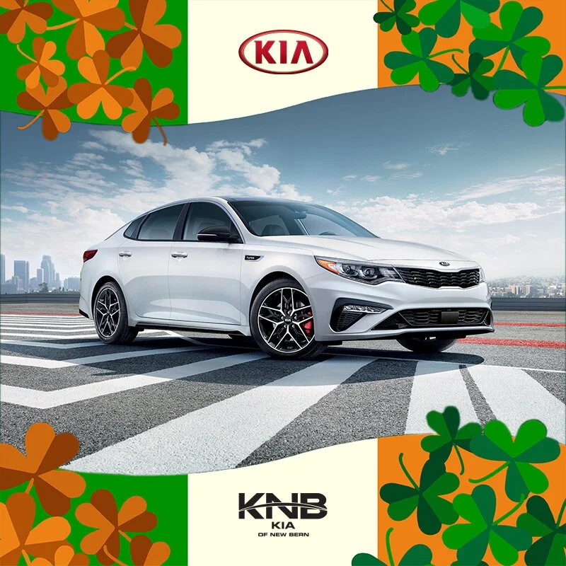 A white Kia sedan parked on a city street with a blue sky and clouds in the background, surrounded by Irish shamrock decorations and Kia branding.