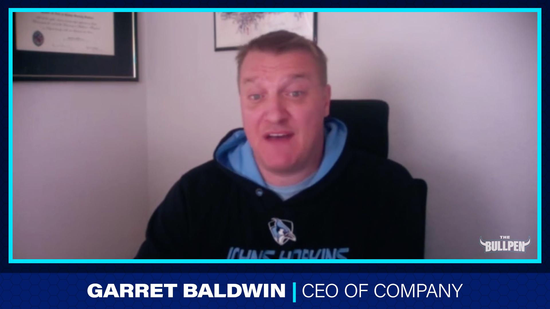 Man sitting in an office chair wearing a black hoodie with a sports team logo, speaking into the camera with a framed certificate and a picture on the wall behind him.