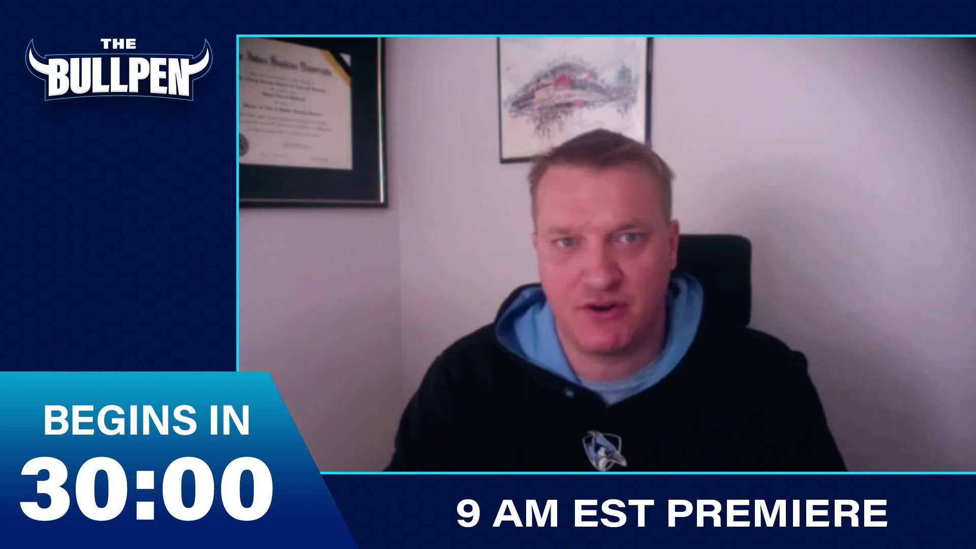 A man wearing a black hockey jersey with a blue hoodie underneath, sitting in front of framed certificates and artwork on a white wall, speaking directly to the camera in a video broadcast