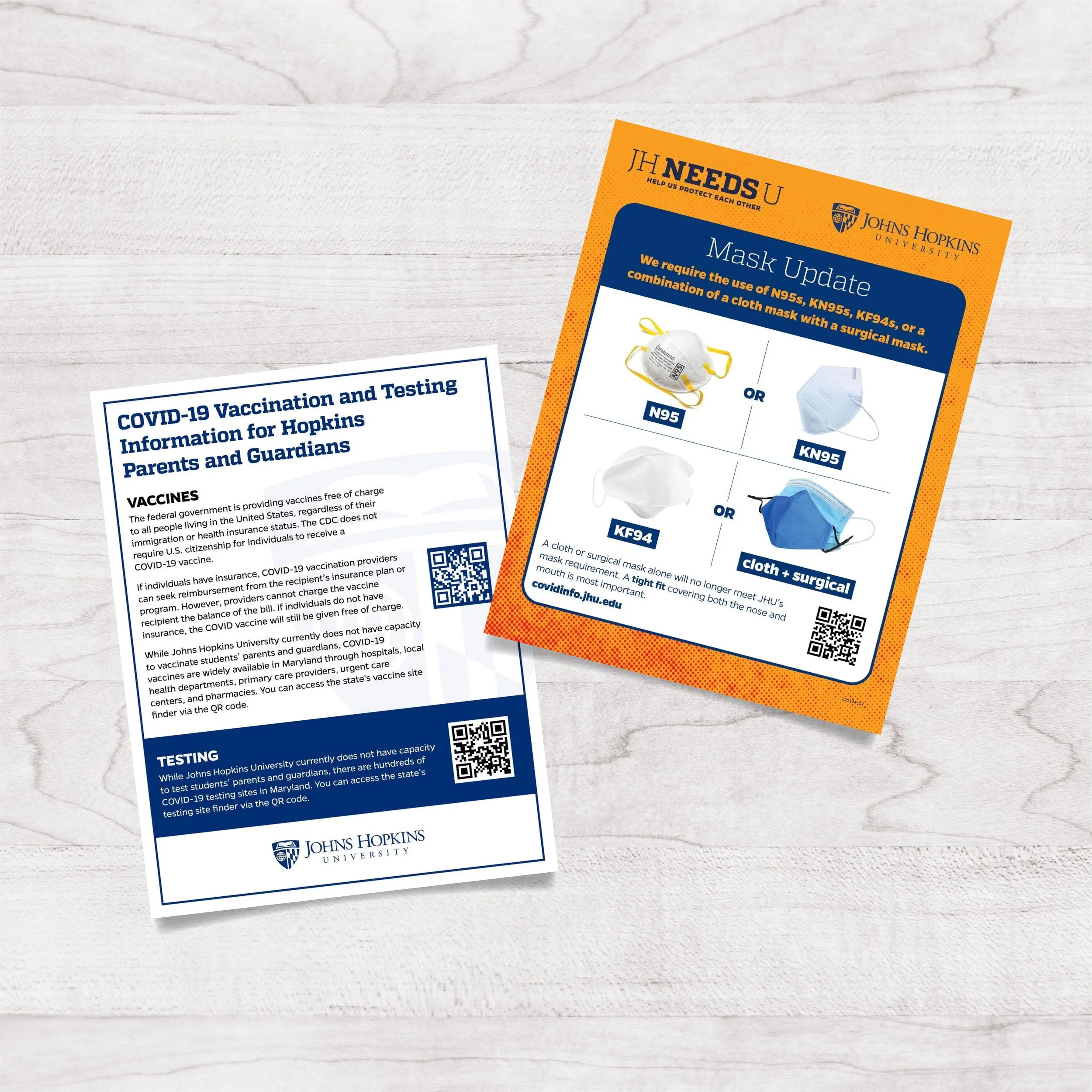 Two COVID-19 informational flyers from Johns Hopkins University on a white textured surface. One flyer discusses vaccination and testing information, the other provides a mask update with images of different types of masks.