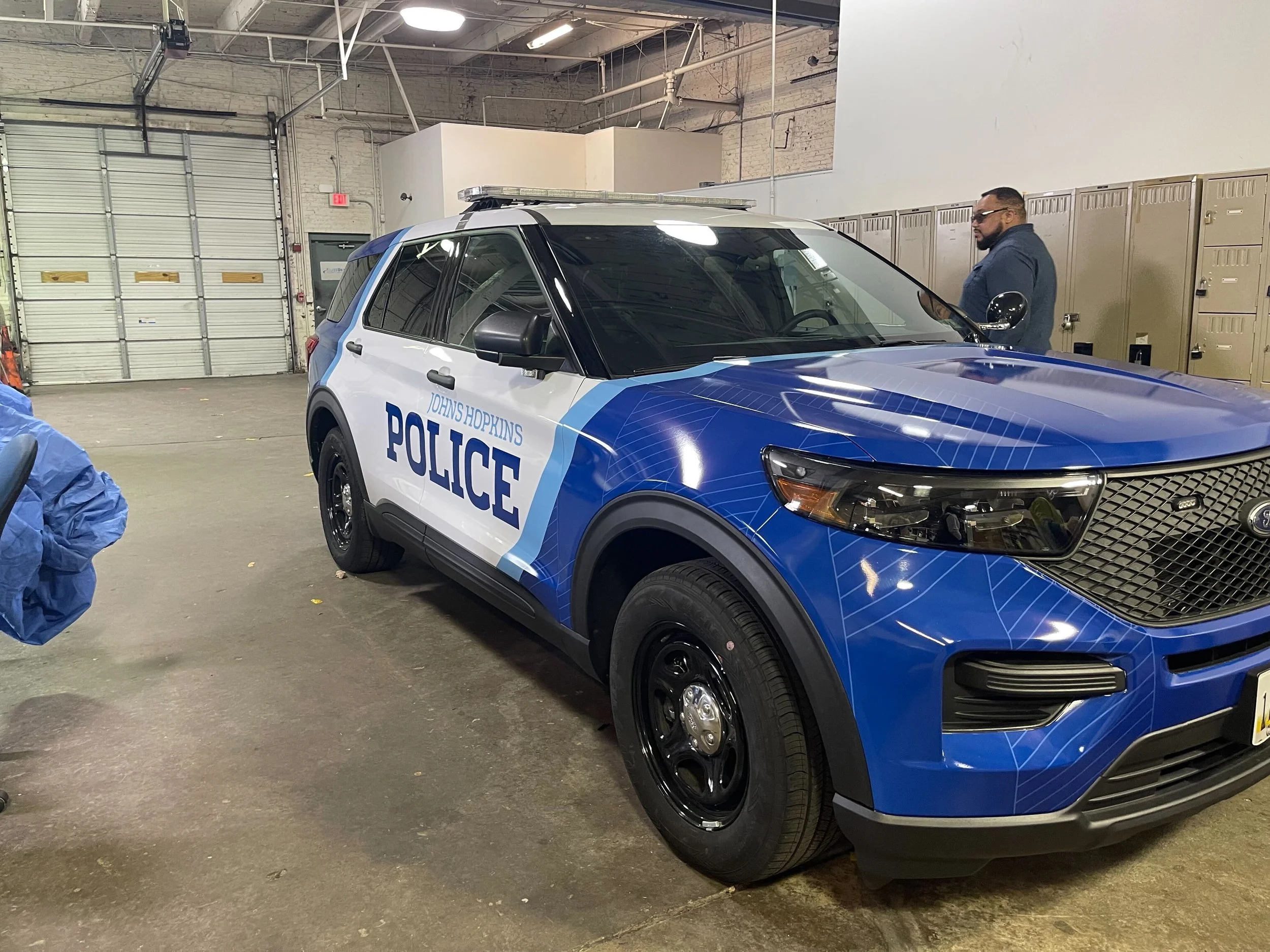 A blue and white police SUV with 'Johns Hopkins POLICE' decals parked inside a garage. A man in a dark jacket is standing near lockers in the background.