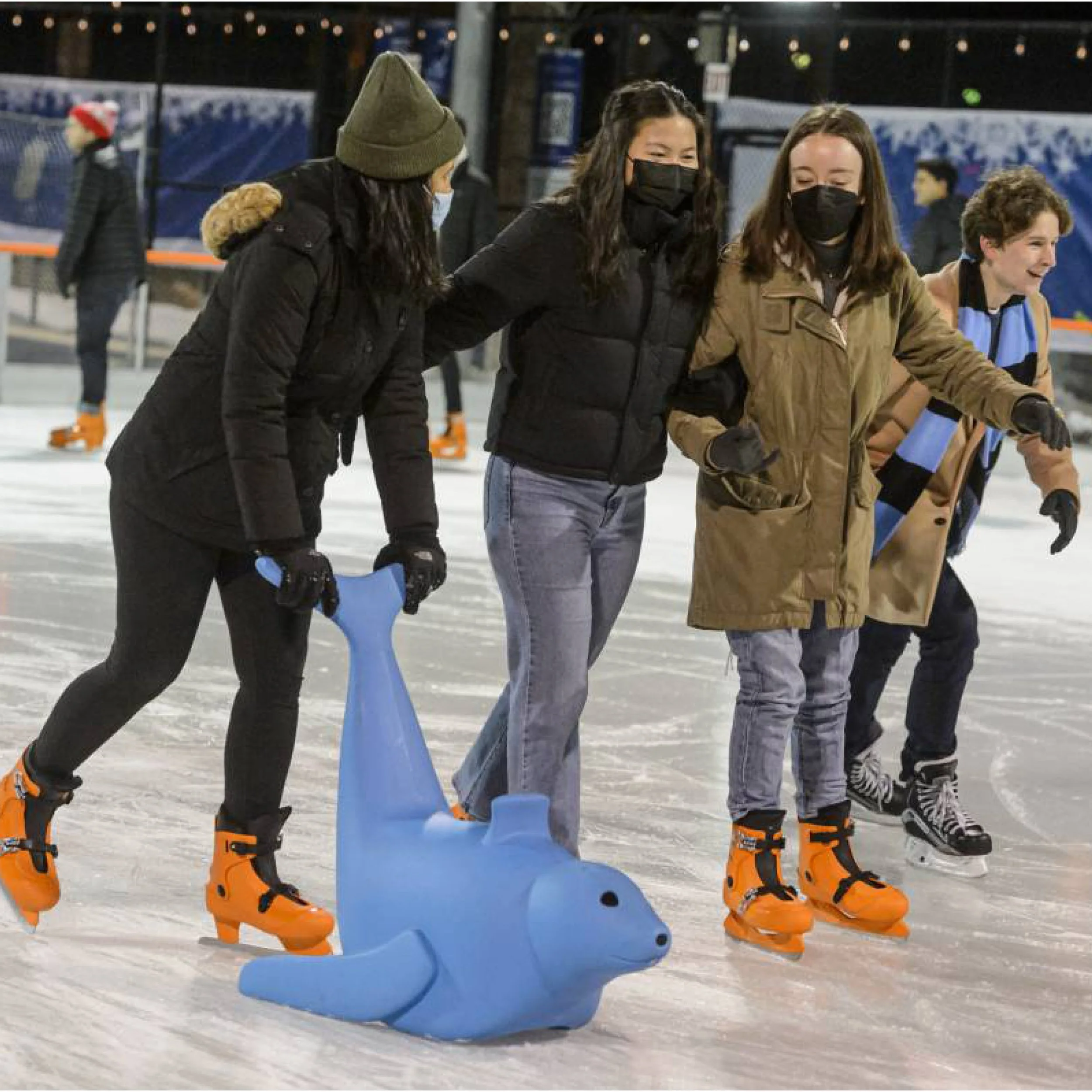 Four people ice skating on an outdoor rink at night, with one person holding and pushing a blue dolphin-shaped skating aid.