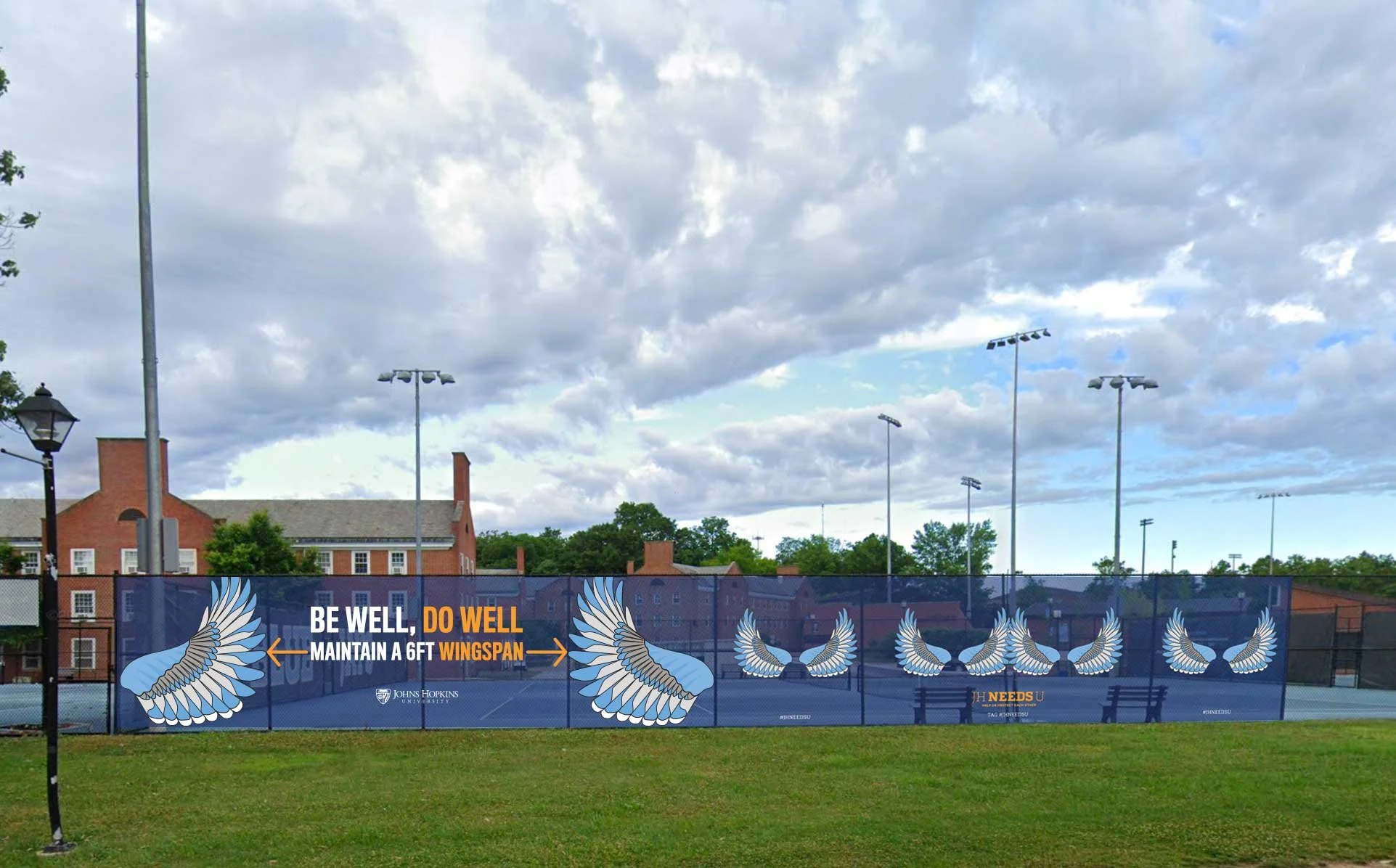 A tennis court with a large banner that reads, 'Be Well, Maintain a 6FT WingSpan' featuring illustrations of wings. The banner promotes health guidelines from Johns Hopkins University. The tennis court is outdoors, with benches, multiple light poles,