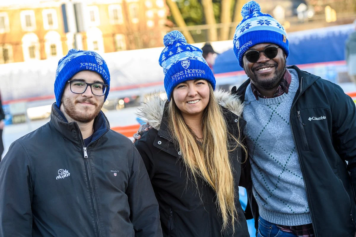 Three people standing outdoors wearing blue knit hats with snowflakes and "Johns Hopkins University" logo, smiling at the camera.
