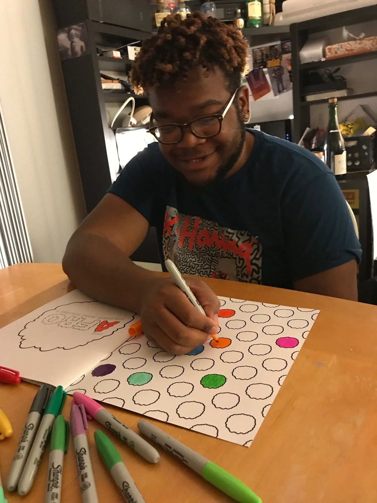 A person with glasses and short curly hair coloring a large sheet of paper with circles in various colors using markers. They are smiling and seem engaged in their activity. The background shows a black bookshelf with various items and bottles.