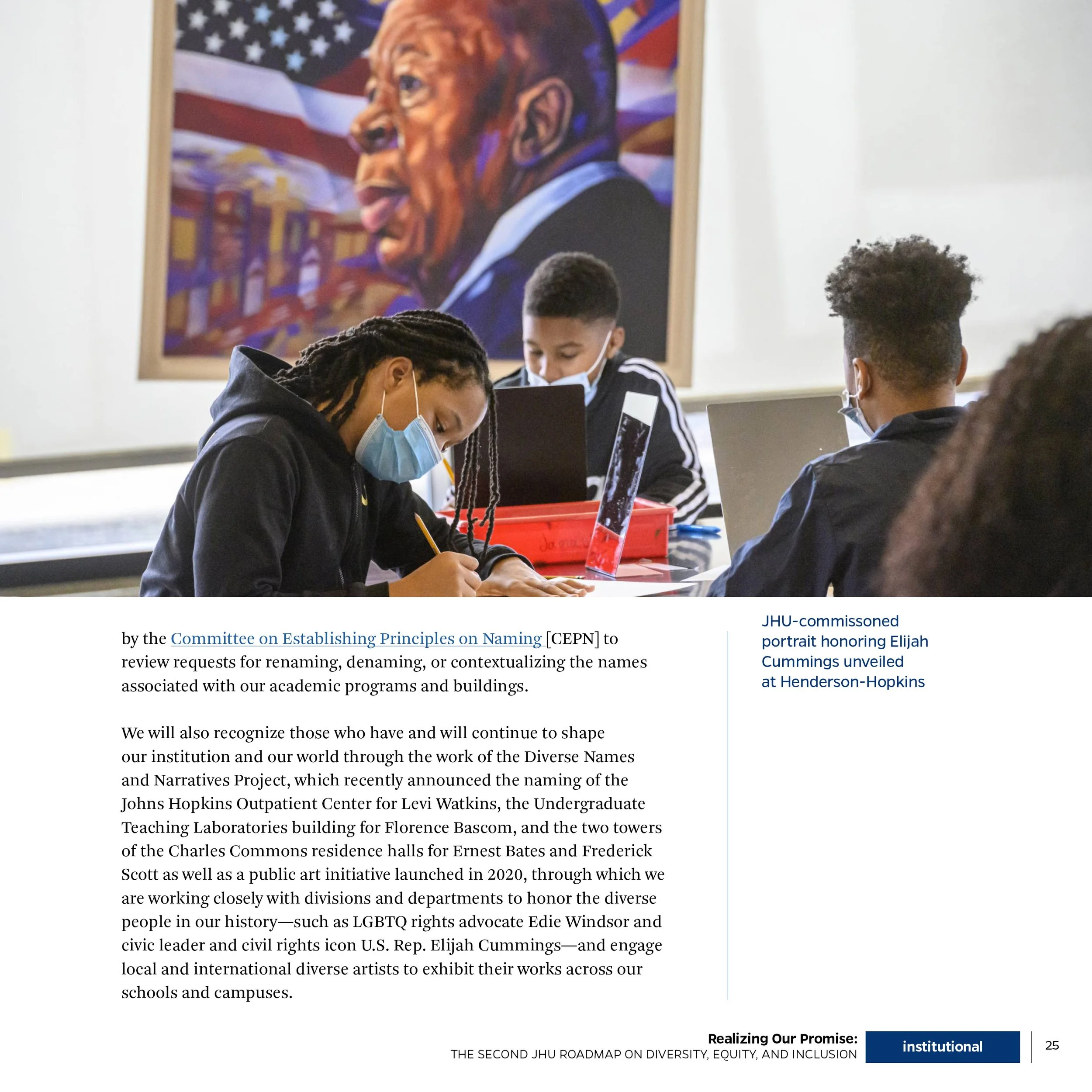 Students seated at desks in classroom, wearing face masks, working on tablets and laptops. A large colorful portrait of a man sticking out his tongue is hanging on the wall behind them.