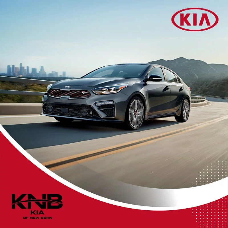 A black Kia sedan driving on a mountain road with a city skyline and mountains in the background, with Kia logos in the top right and bottom left corners.
