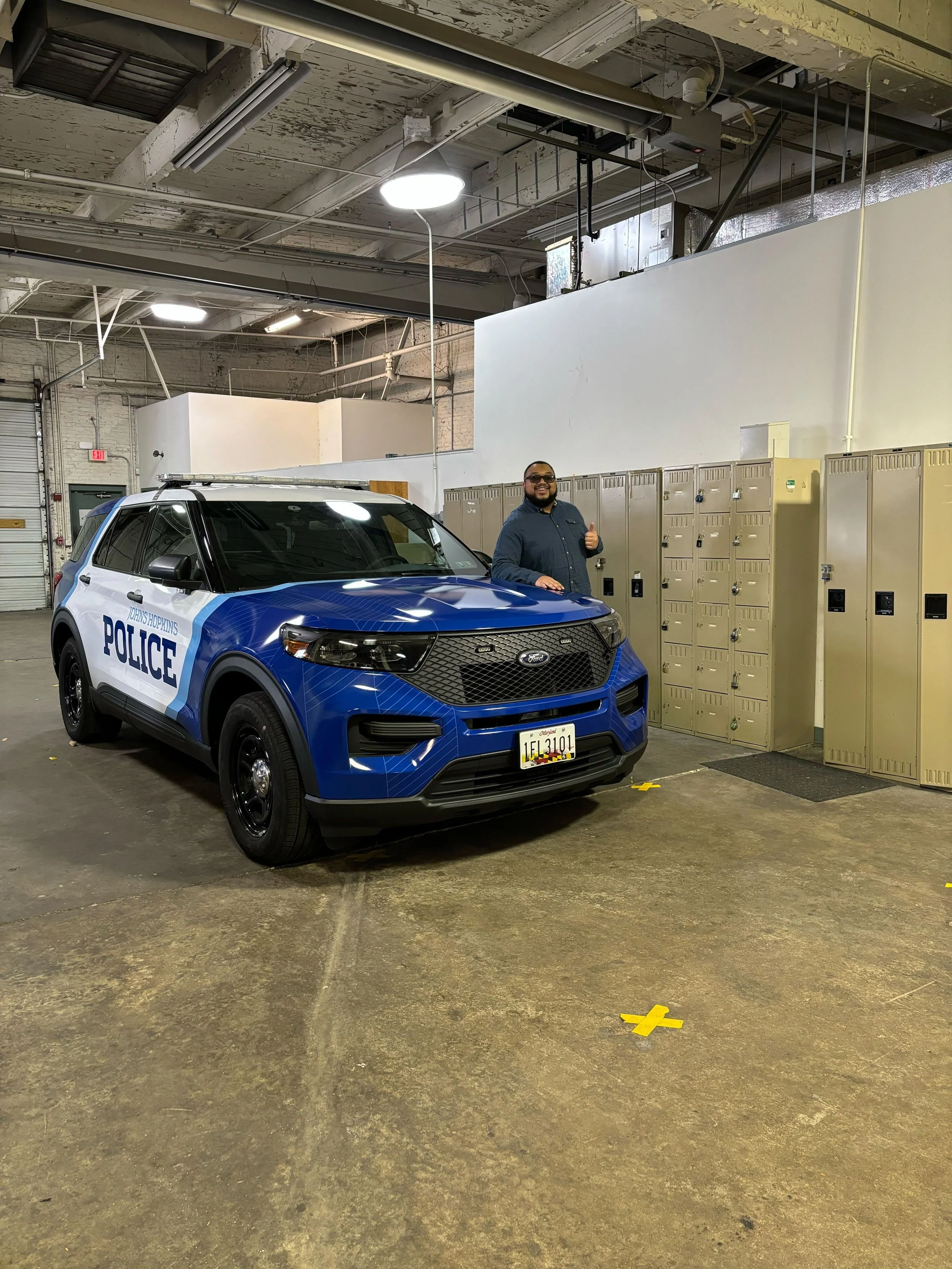A man standing next to a blue and white police vehicle in a garage with lockers and industrial ceiling lighting.