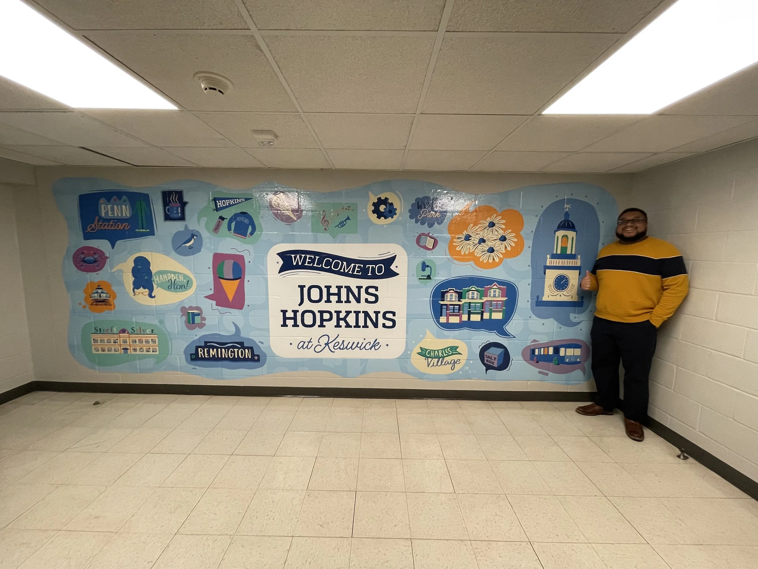 A man standing next to a wall mural that reads 'Welcome to Johns Hopkins at Keswick.' The mural has illustrations of buildings, a clock tower, a bus, and various decorative elements.