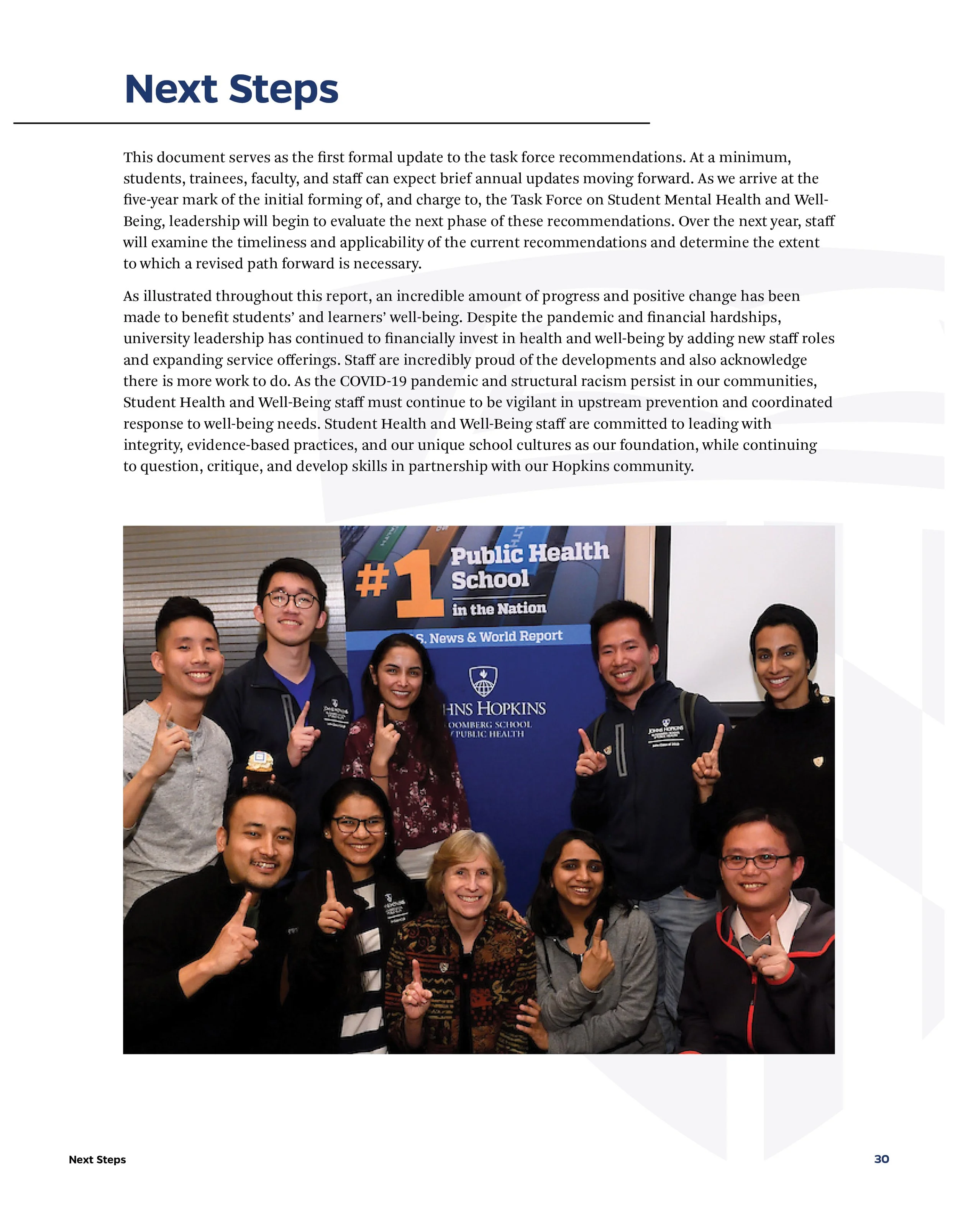 Group of diverse people standing in front of a sign that reads '#1 Public Health School in the Nation' at Johns Hopkins. They are smiling and holding up one finger.