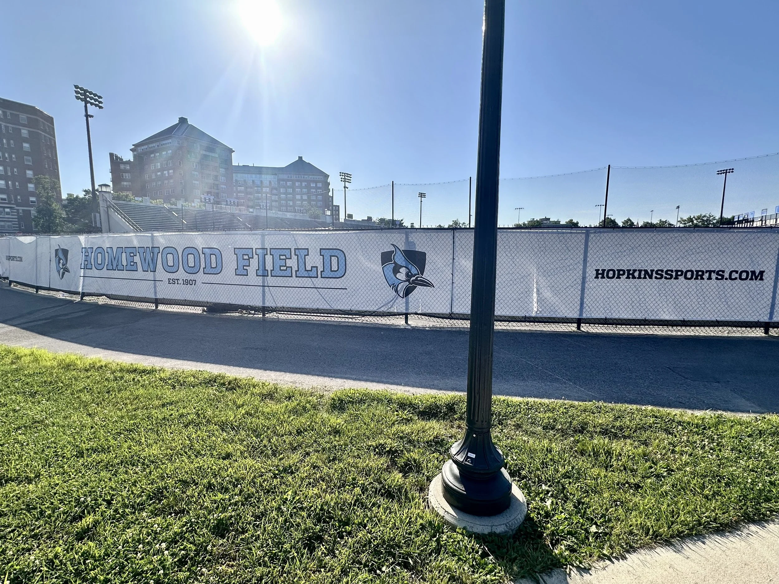 A sports field with a chain-link fence and a banner reading 'Homewood Field' with a school or stadium complex and tall light poles in the background, under a clear blue sky.
