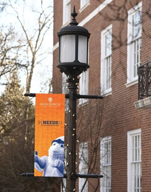 A black vintage-style street lamp with decorative top and four crossbars, adorned with a Johns Hopkins banner and string lights, situated in front of a brick building with white window frames and a small balcony.