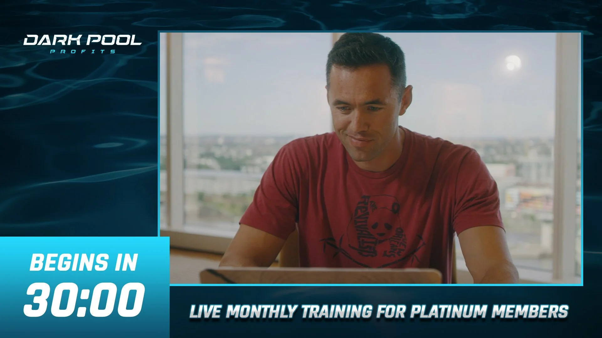 A man wearing a red T-shirt looking at a laptop in a bright room with a cityscape view outside the window.