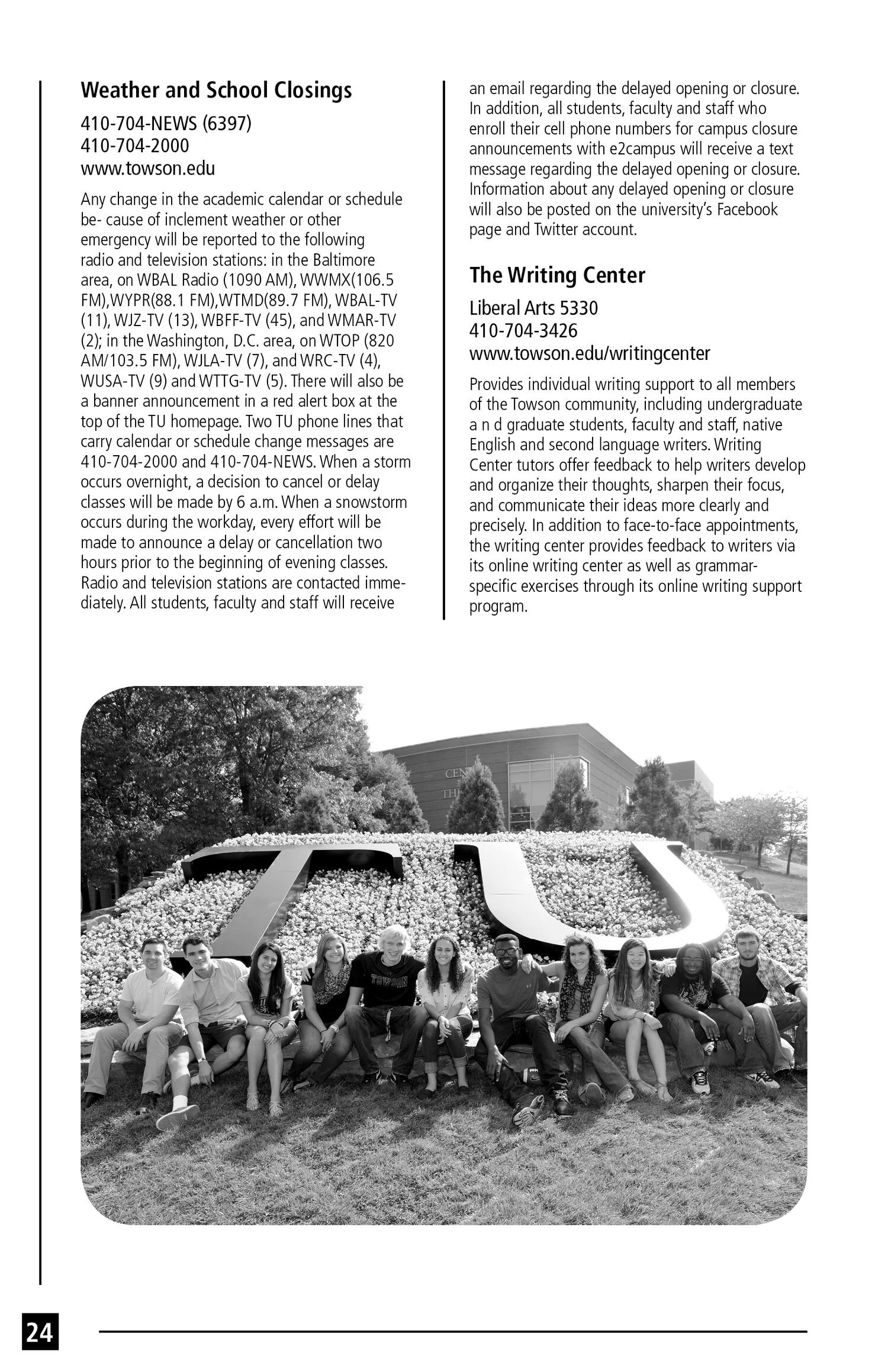 A black-and-white photo of a group of students sitting in front of a university sign surrounded by flowers, with a campus building and trees in the background.