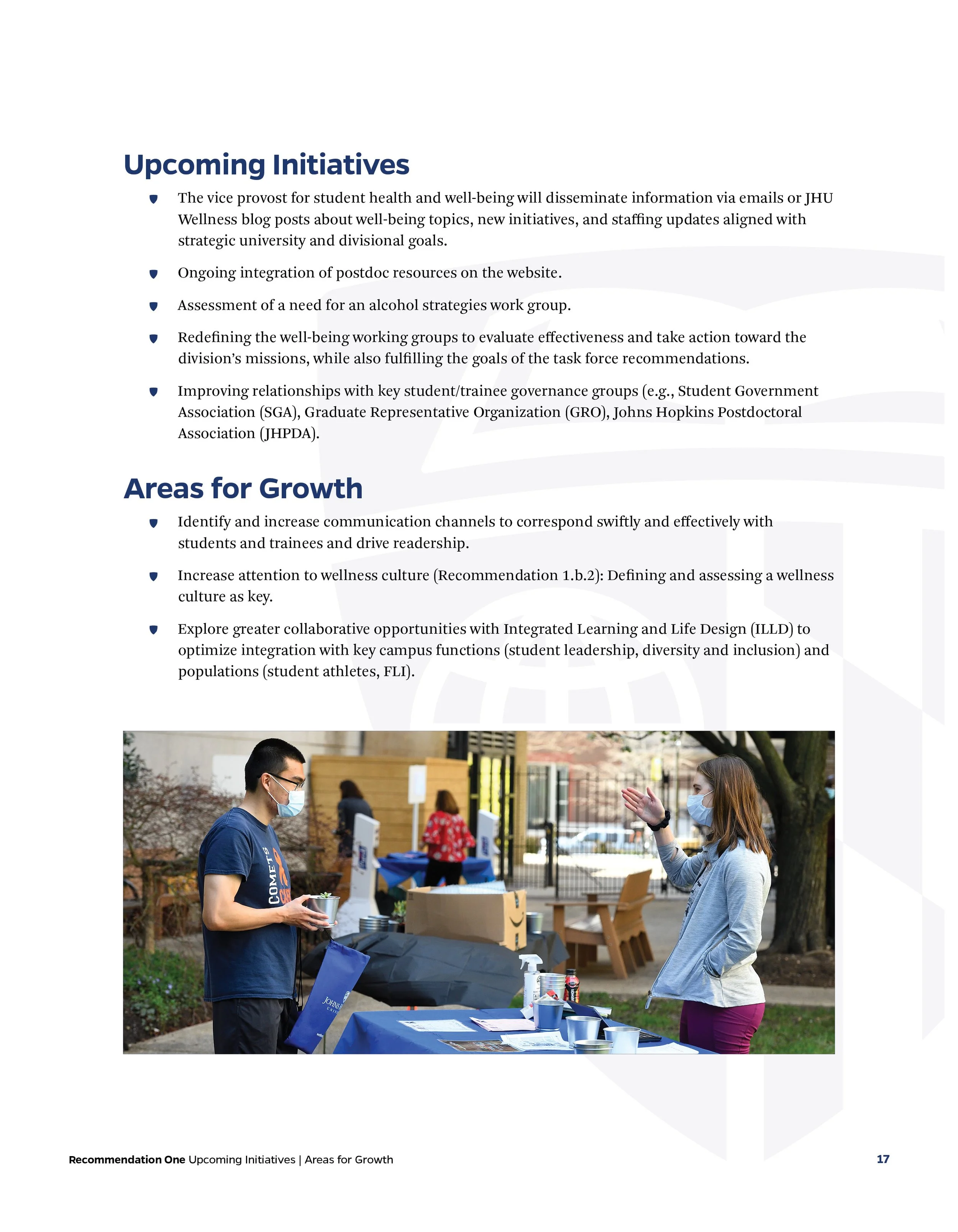 Two people wearing face masks engage in conversation outdoors at a table with informational materials, with other tables and people visible in the background.