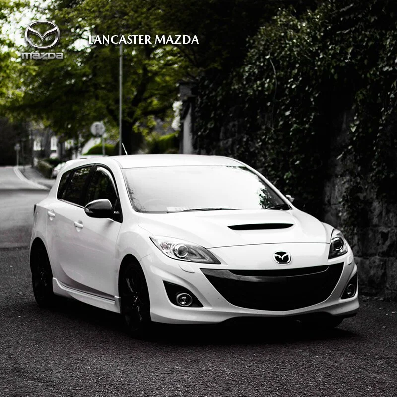 A white Mazda sedan parked on a street with greenery and trees in the background. The image features Lancaster Mazda branding at the top.