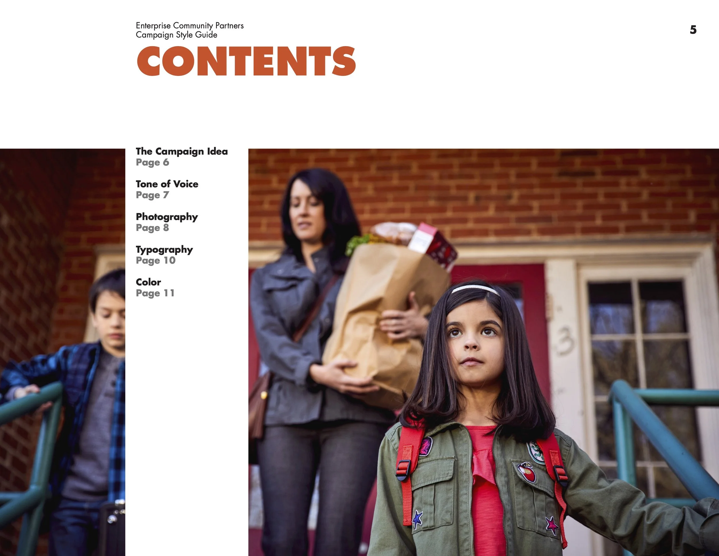 A woman with dark hair holding a paper bag of groceries, standing in front of a red brick house with two children. One girl with long dark hair and a green jacket with patches looks forward, while a boy with short dark hair, a blue plaid shirt, and a