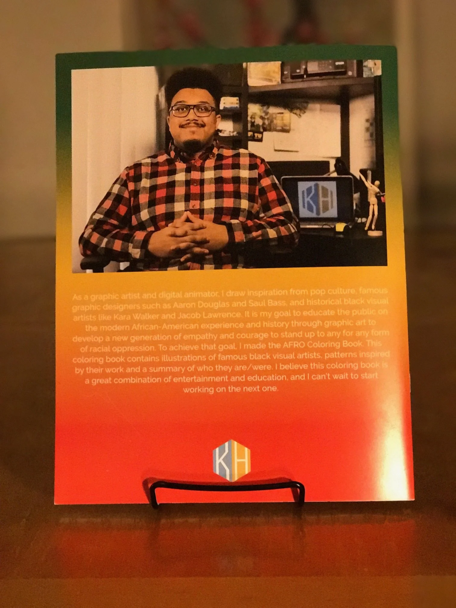 Back cover of a coloring book featuring a photo of a man with black hair, glasses, and a colorful plaid shirt, sitting at a desk with a computer and art supplies in a room with shelves.