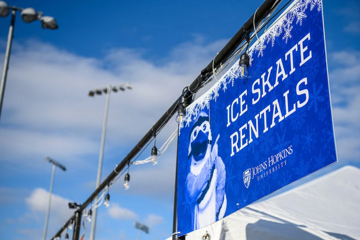 Blue sign displaying 'Ice Skate Rentals' with the Johns Hopkins University logo, featuring a mascot, under a partly cloudy sky with stadium lights in the background.