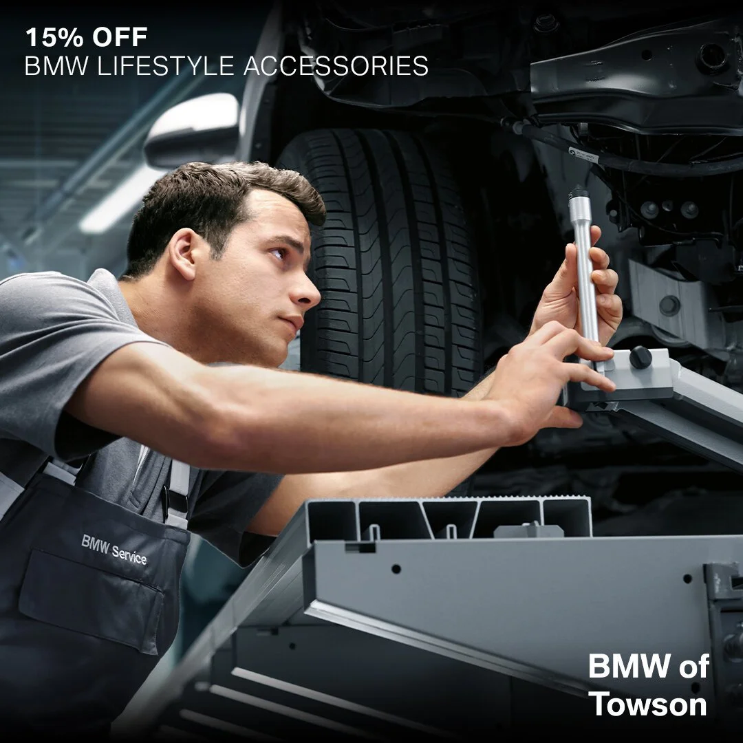 A mechanic working on a car's undercarriage in a service shop, wearing a BMW Service apron, with a focus on the tire and tools.