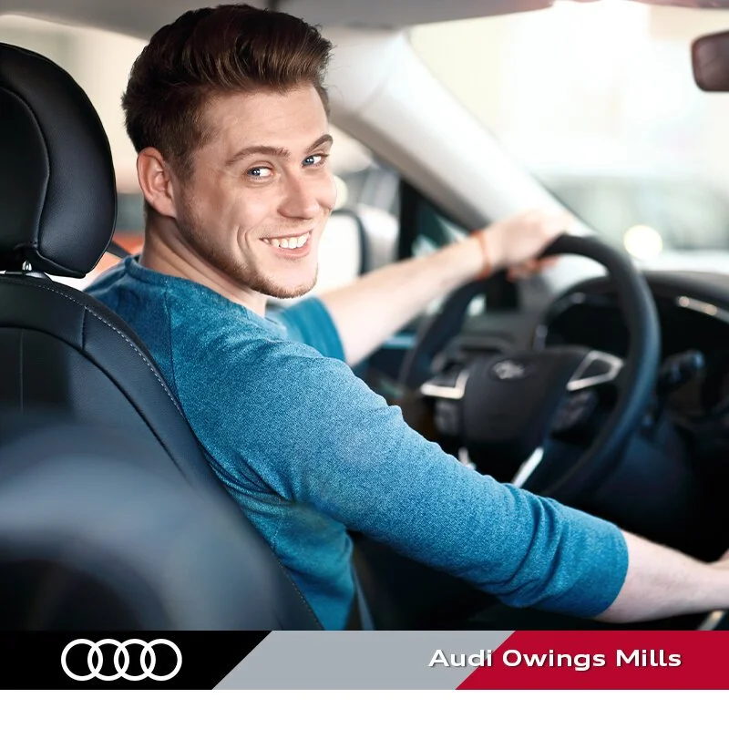 A young man sitting in the driver's seat of a car, smiling and looking back at the camera, wearing a blue shirt, inside a modern vehicle.