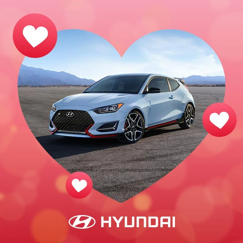 A light blue Hyundai car with black accents on a large, open, outdoor paved area with mountains in the background, framed by a pink heart-shaped overlay with heart icons and the Hyundai logo and name.