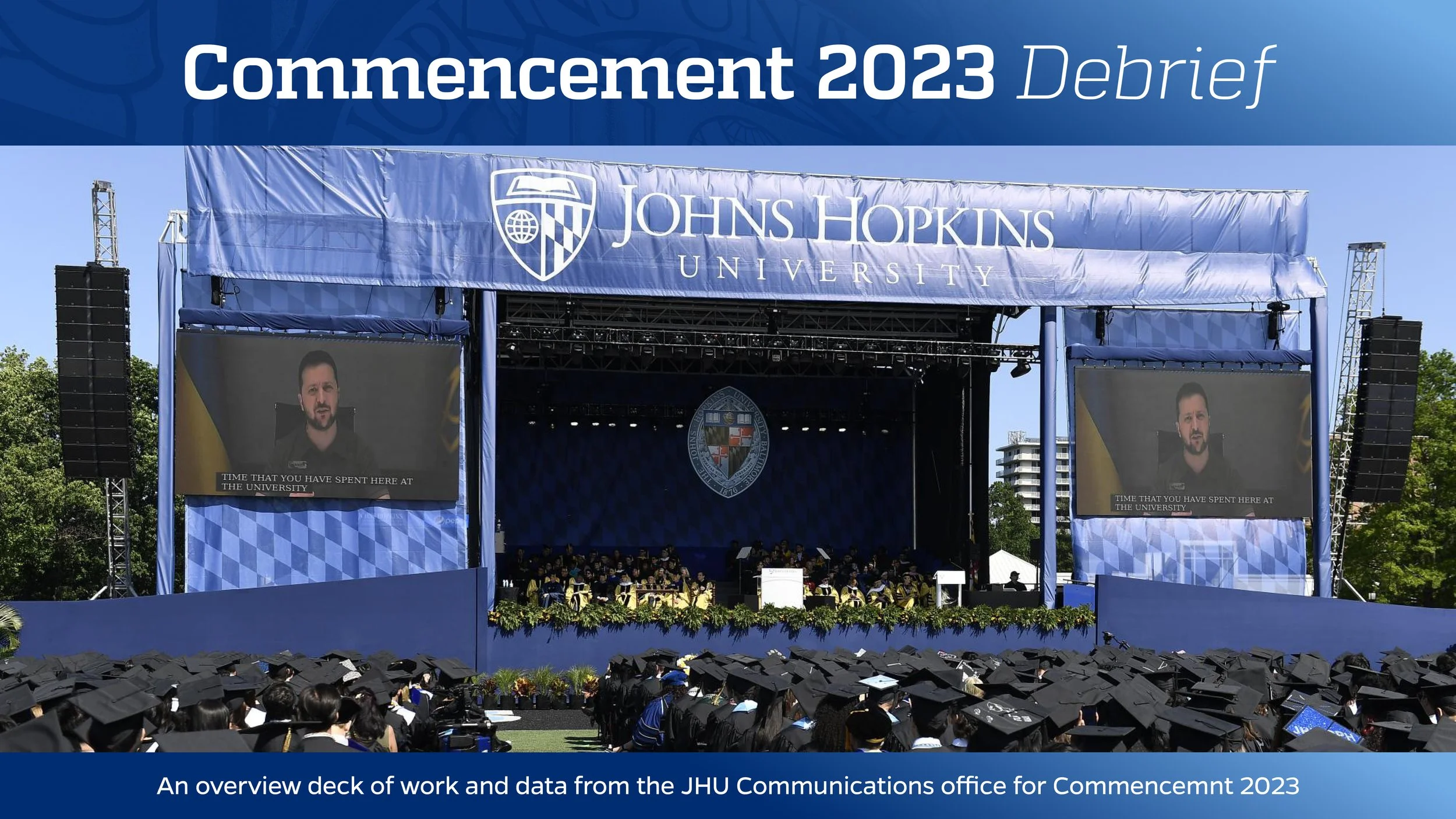 Stage at Johns Hopkins University commencement ceremony 2023 with graduates in caps and gowns, large screens showing a speaker, and a banner overhead reading 'Commencement 2023 Debrief'.