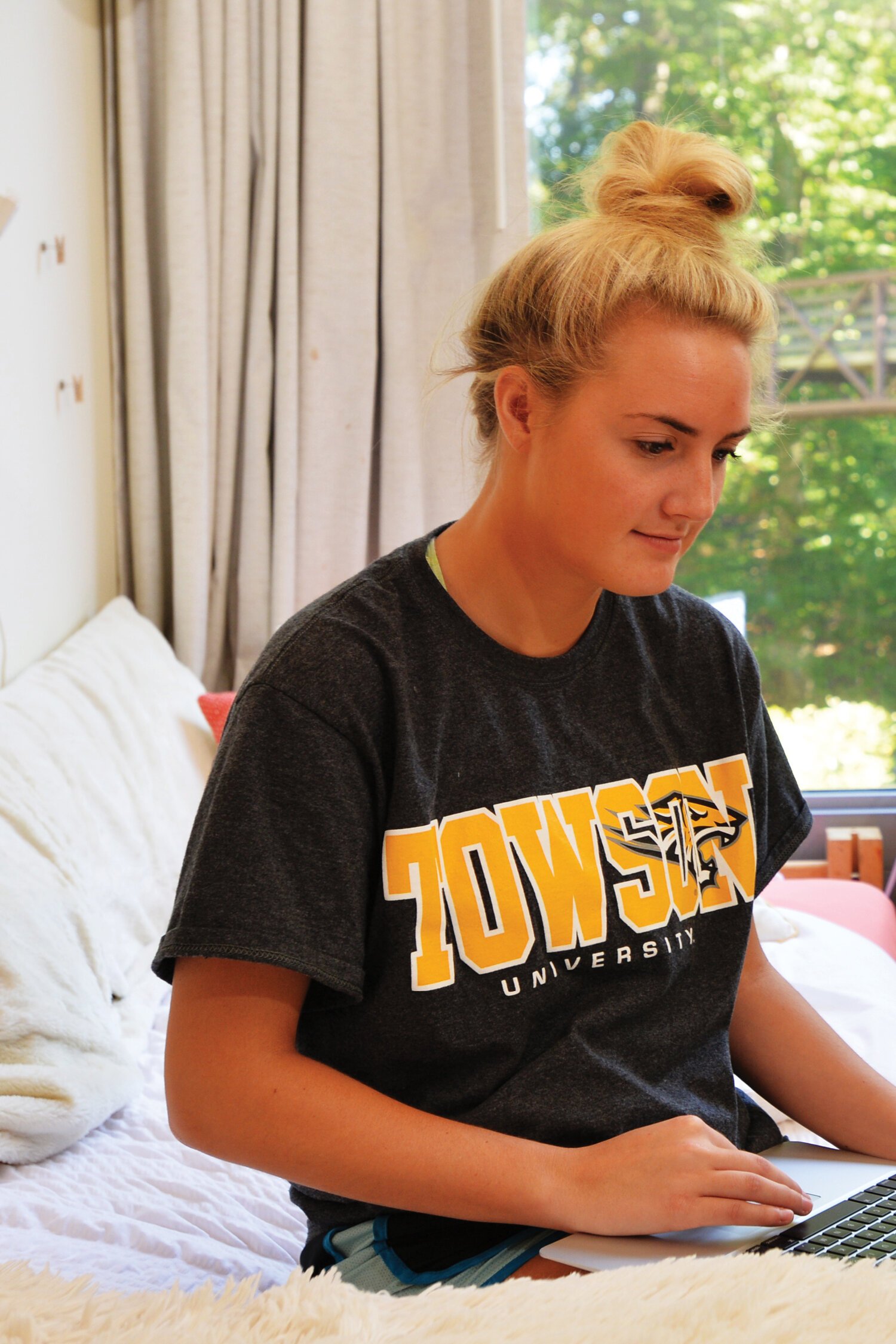 A young woman with blonde hair tied in a bun, wearing a black Towson University t-shirt, sitting on a bed using a laptop in a bright room with large windows and green trees outside.